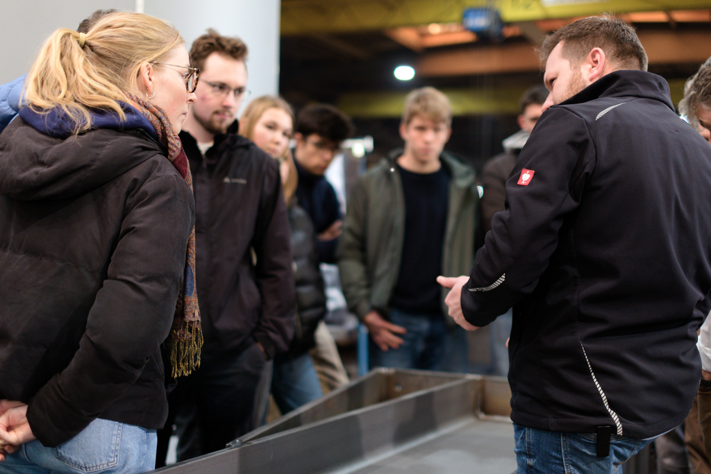 Gruppe junger Erwachsener lauscht einer Person im Vordergrund, die etwas erklärt; Innenraum, informelles Treffen.